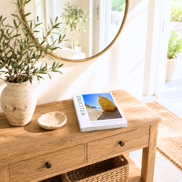 A styled interior scene showing a light wood console table with a woven basket underneath, a round gold-framed mirror on the wall, a potted olive branch in a textured cream vase, a small dish, and a airbnb welcome book on top.