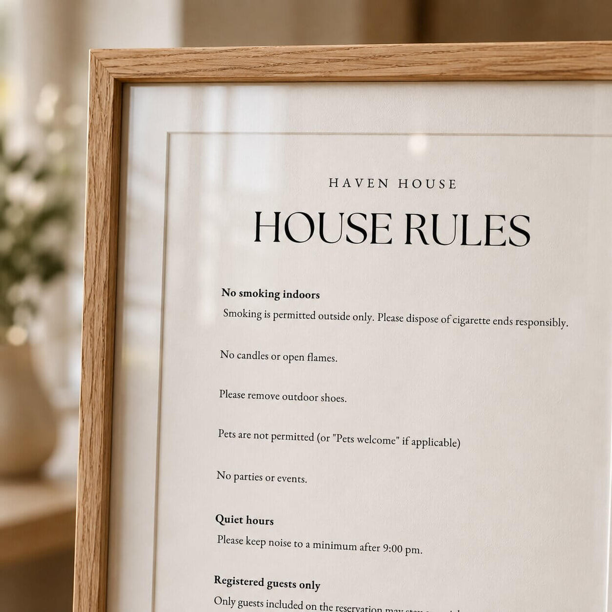 A framed 'Haven House House Rules' sign with black text on a white background, enclosed in a light natural wood frame. The blurred background shows a bright, elegant indoor setting with soft lighting and a hint of greenery.