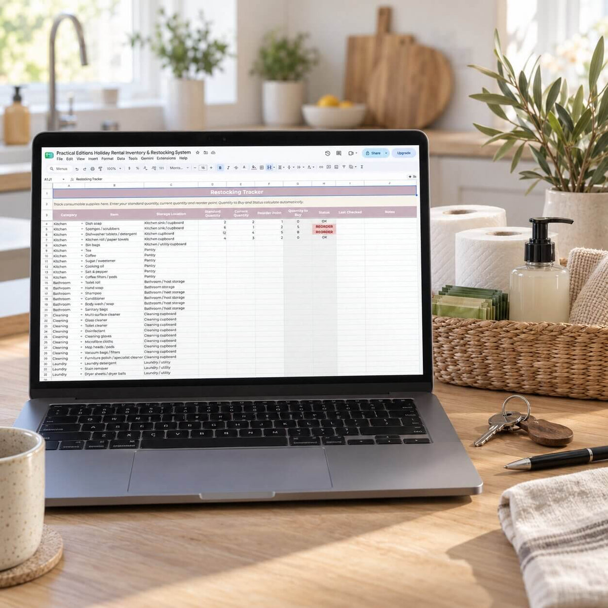 A laptop on a wooden kitchen counter displaying a spreadsheet template titled 'holiday rental restocking tracker'. The scene is styled with a coffee mug, keys, a pen, a basket, and kitchen plants in the background.