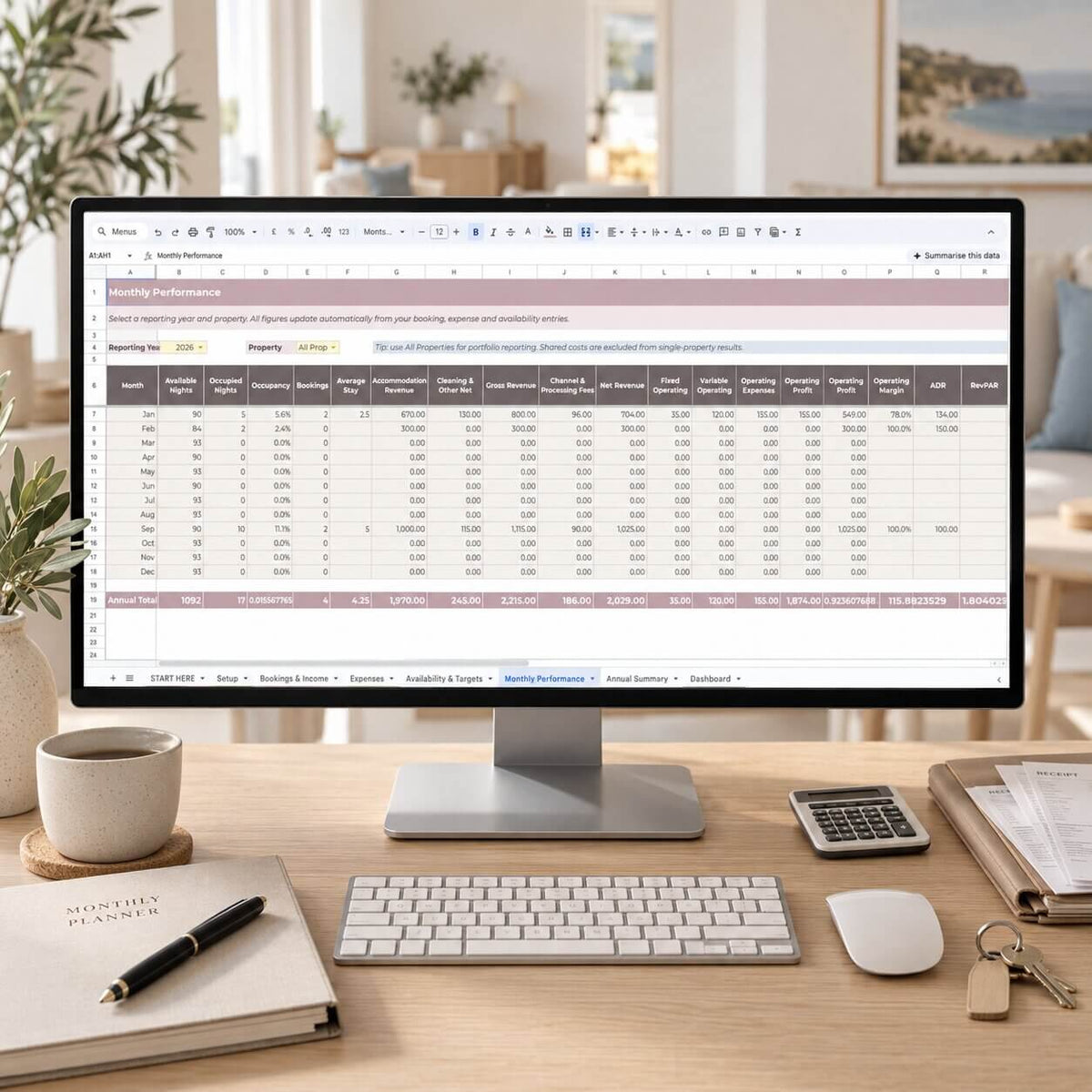 A desktop computer monitor on a wooden desk displaying a spreadsheet template, surrounded by a keyboard, mouse, calculator, coffee cup, notebook, and keys in a bright home office setting.