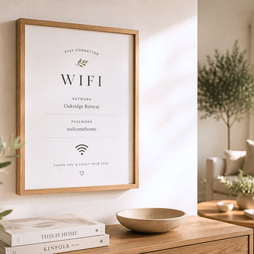 A framed minimalist WiFi sign with black text on a white background hangs on a light wall in a styled interior. The sign includes the text 'STAY CONNECTED', 'WIFI', 'NETWORK Oakridge Retreat', 'PASSWORD welcomehome', and 'THANK YOU & ENJOY YOUR STAY'.