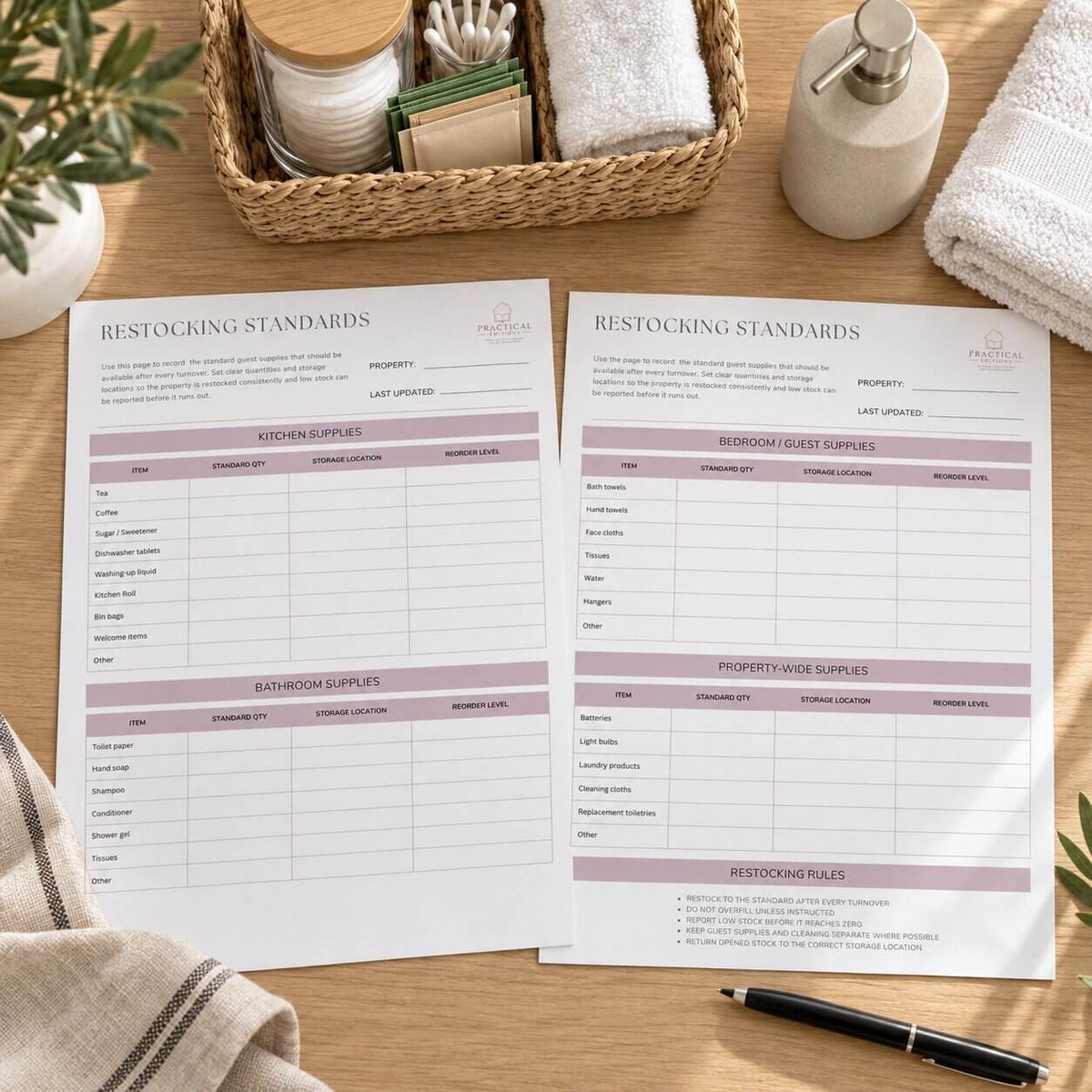 Two printed Restocking Standards sheets laid on a wooden table, surrounded by towels, a soap dispenser, a basket of cotton supplies, and a pen. The sheets contain tables for kitchen, bathroom, and property-wide supplies with lavender header bands.