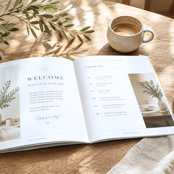 An open printed magazine or book laid on a wooden table, showing a welcome page and contents layout with soft interior design photos. A ceramic mug of coffee sits nearby, along with leafy branches and a draped fabric, in a warm styled setting.